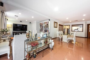 A spacious living room with a flat-screen TV, a bookshelf, a dining area with white chairs, and a glass coffee table. - Hillside City-View Retreat with Pool & Alfresco, Near City (Dianella)
