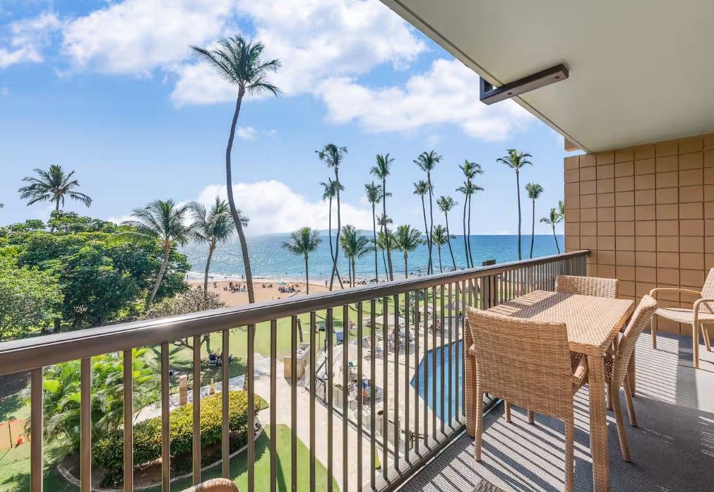 A balcony with wicker furniture overlooking a beach with palm trees and a pool.