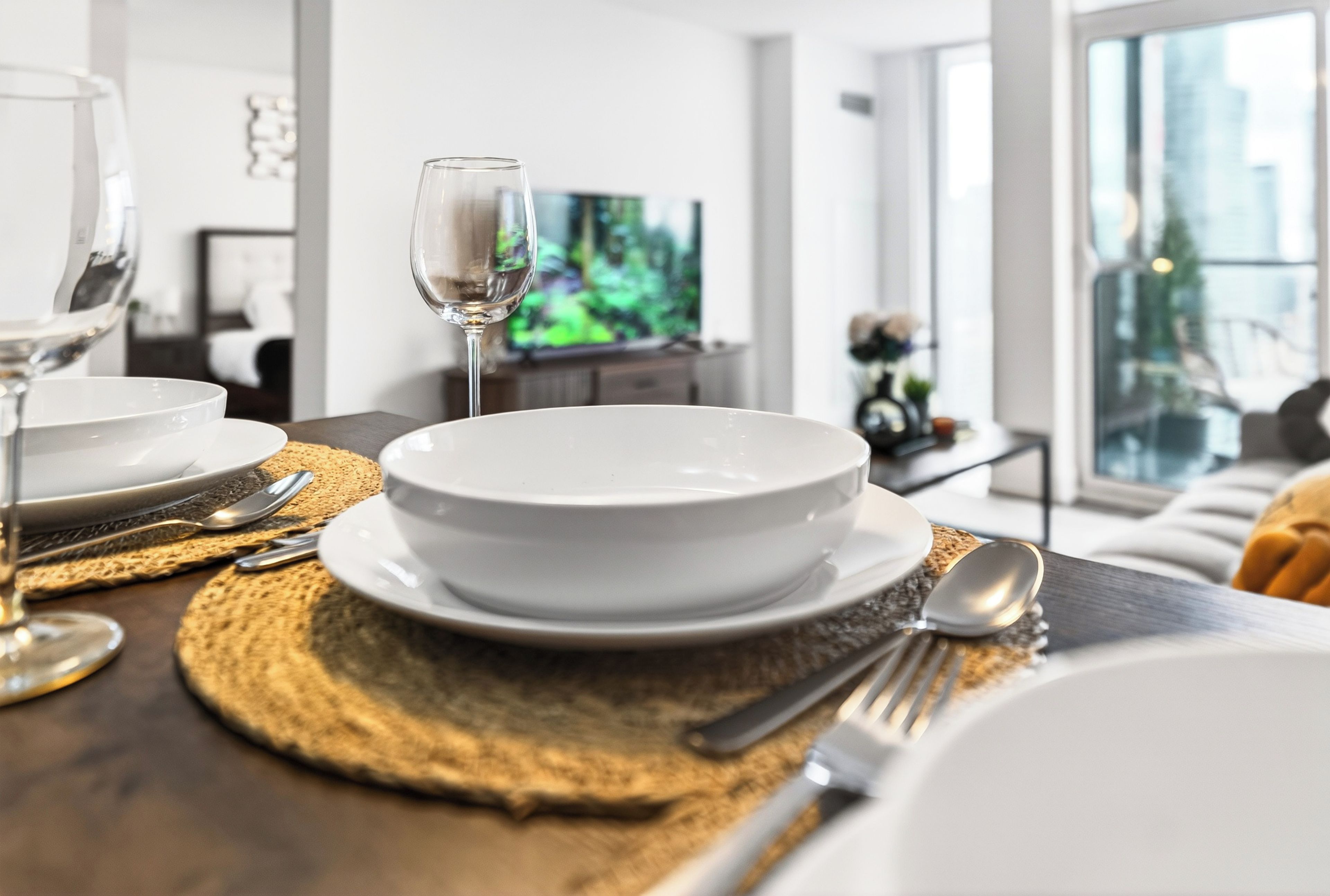 Una mesa de comedor dispuesta con platos, fuentes y cubiertos blancos, una copa de vino y un mantel individual.