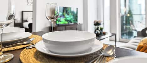 Una mesa de comedor dispuesta con platos, fuentes y cubiertos blancos, una copa de vino y un mantel individual.