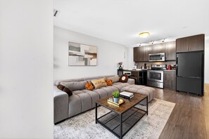 Ein modernes Wohnzimmer mit Ecksofa, Couchtisch und Küchenzeile, ausgestattet mit dunklen Schränken, einer Mikrowelle und einem Kühlschrank.