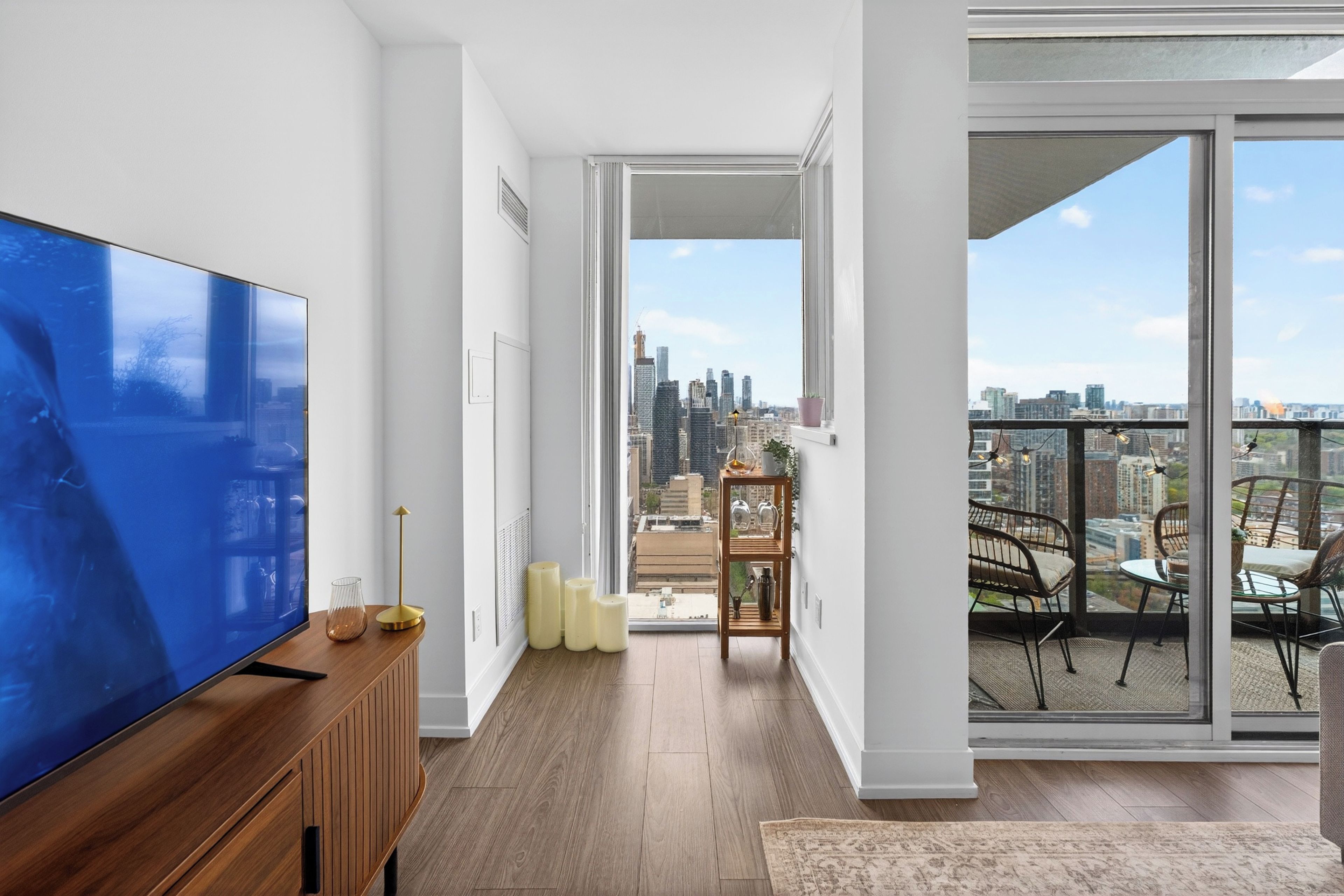 A modern living room with a large flat-screen TV, a wooden console, and a view of the city skyline through a large sliding glass door.