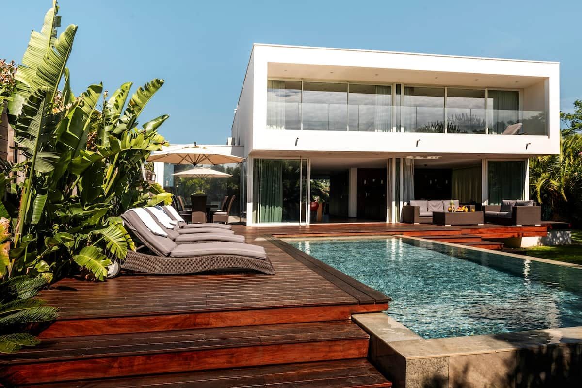 A modern two-story house with a swimming pool, lounge chairs, and tropical plants.