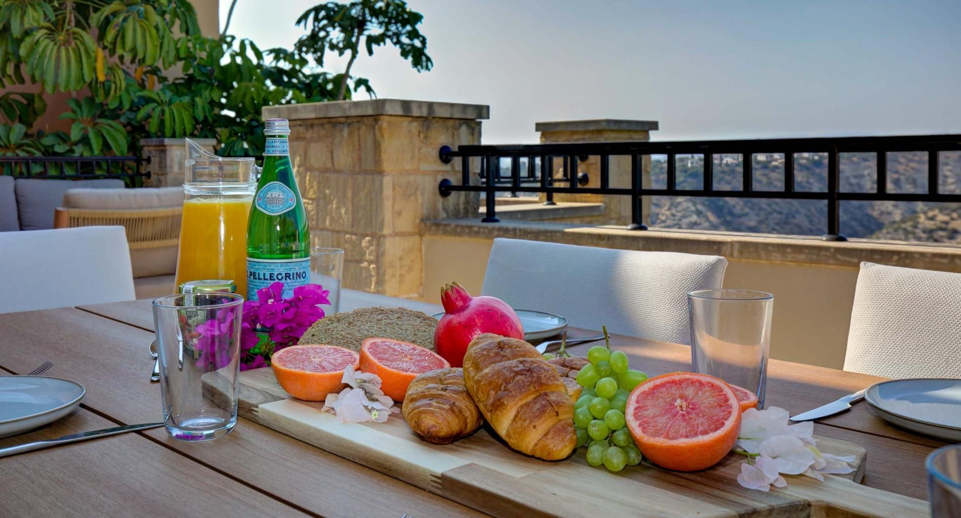 A table set with breakfast items including bread, fruits, and a beverage, with a view of the landscape in the background.