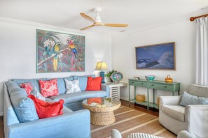 Ein Wohnzimmer mit einem blauen Sofa, roten und weißen Kissen, einem gewebten Couchtisch, einem grünen Beistelltisch und einem Vogelbild.