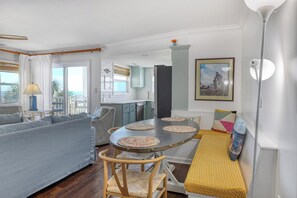 Un salón acogedor con un sofá, una mesa redonda con individuales, un banco con cojines y vistas al océano a través de amplias ventanas.