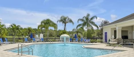 Piscina con un elemento acuático en el centro, rodeada de sillas de descanso y palmeras.