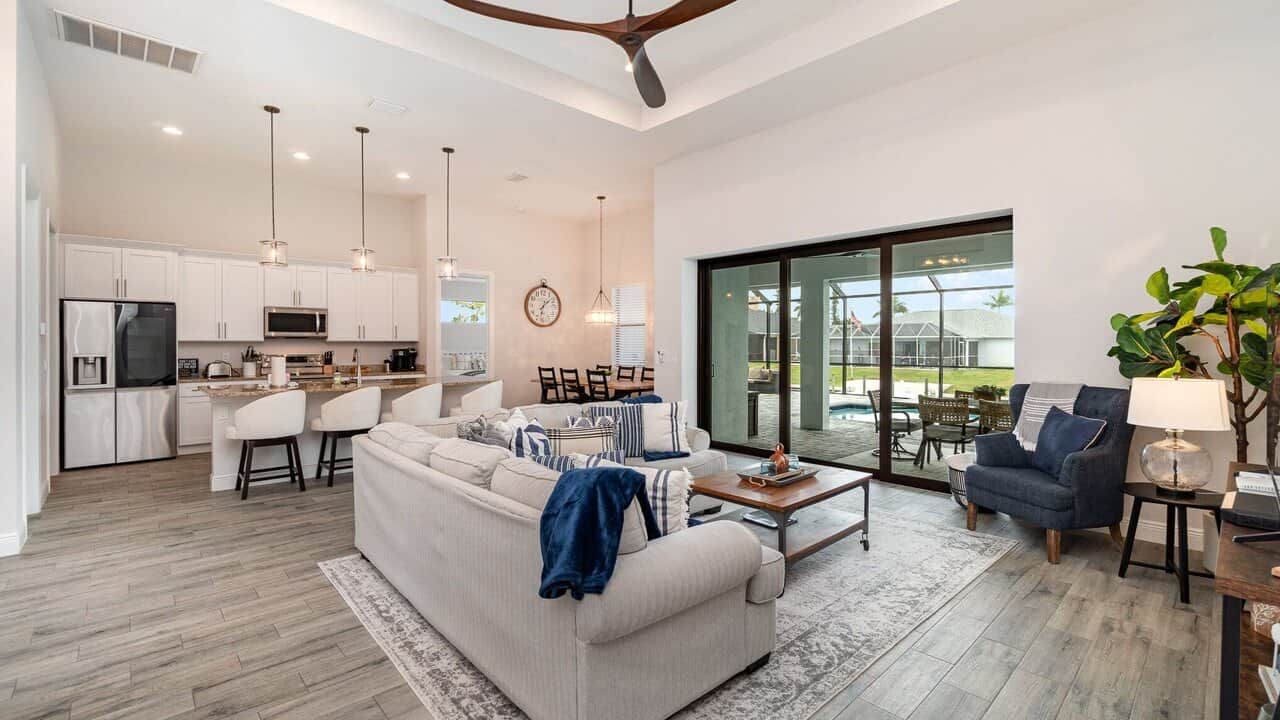 A modern kitchen with white cabinets, a dining area with a table and chairs, a living room with a sofa and a coffee table, and a large sliding glass door leading to an outdoor area.