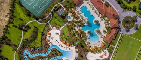 An aerial view of a resort with a large swimming pool, tennis courts, and a winding water slide.