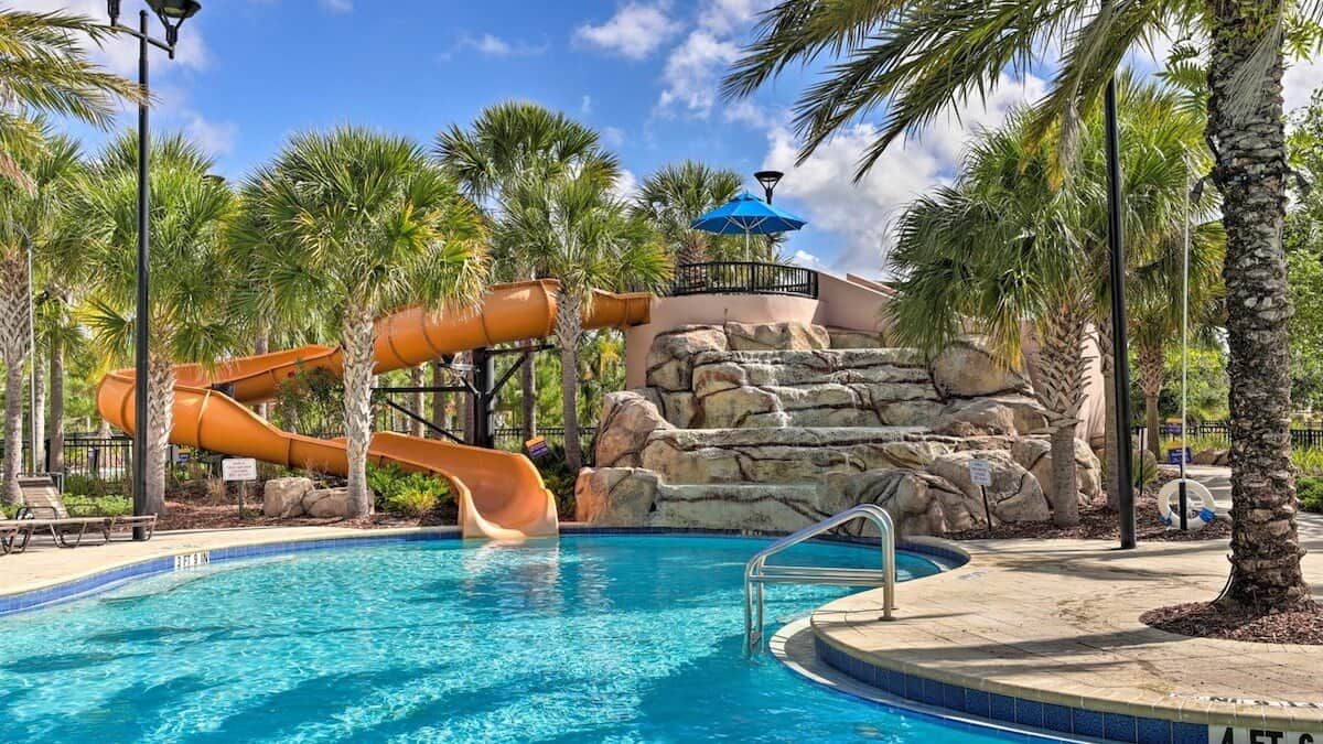 A swimming pool with a water slide, palm trees, and a rocky backdrop.