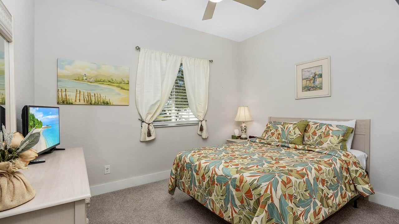 A bedroom with a bed, a window with curtains, a desk with a computer, and artwork on the walls.