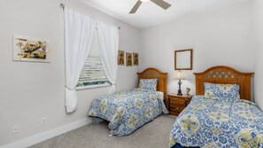 Un dormitorio con dos camas individuales, un ventilador de techo, una ventana con cortinas blancas y obras de arte colgadas en la pared.