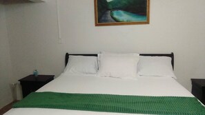 Un llit amb roba de llit blanca i una manta verda, una tauleta de nit amb una làmpada, i un quadre a la paret.