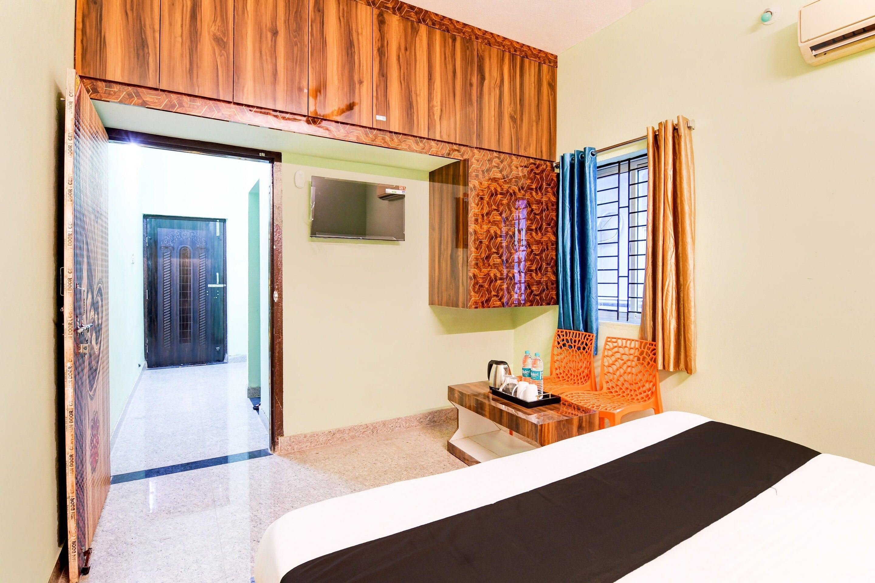 A hotel room with a bed, a bedside table with a kettle and water bottles, a mirror, and a window with curtains.
