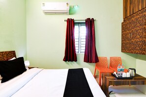 Una habitación de hotel con una cama, una mesita de noche, una mesa con hervidor y botellas de agua, y un aire acondicionado instalado en la pared.