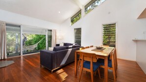 A modern living room with a sofa, dining table, and chairs, large windows with a view of greenery, and a hammock outside.