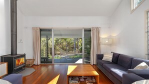 A modern living room with a fireplace, a sofa, a coffee table, and a view of a hammock and trees through large glass doors.