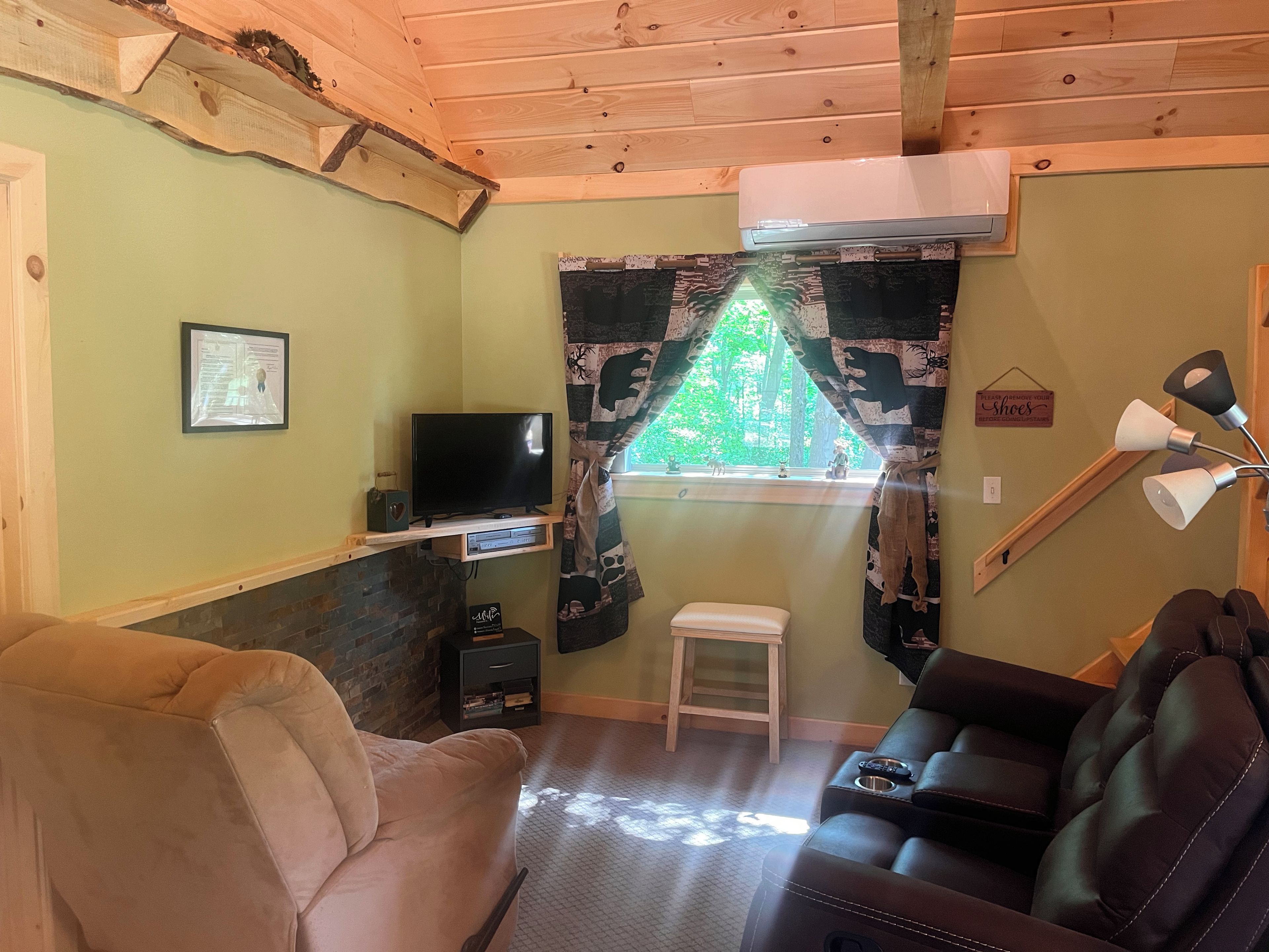 Una habitación acogedora con televisión, un taburete pequeño, un sofá y una ventana que da a vistas de árboles.