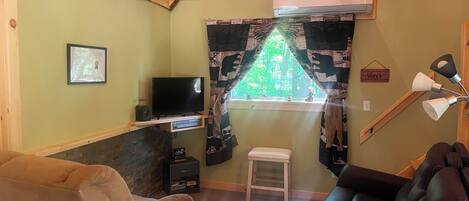 Una habitación acogedora con televisión, un taburete pequeño, un sofá y una ventana que da a vistas de árboles.