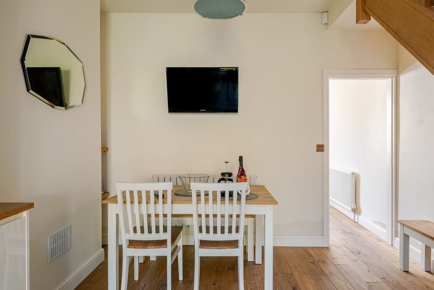 A dining area with a wooden table and white chairs, a flat-screen TV mounted on the wall, and a view of a hallway leading to another room.