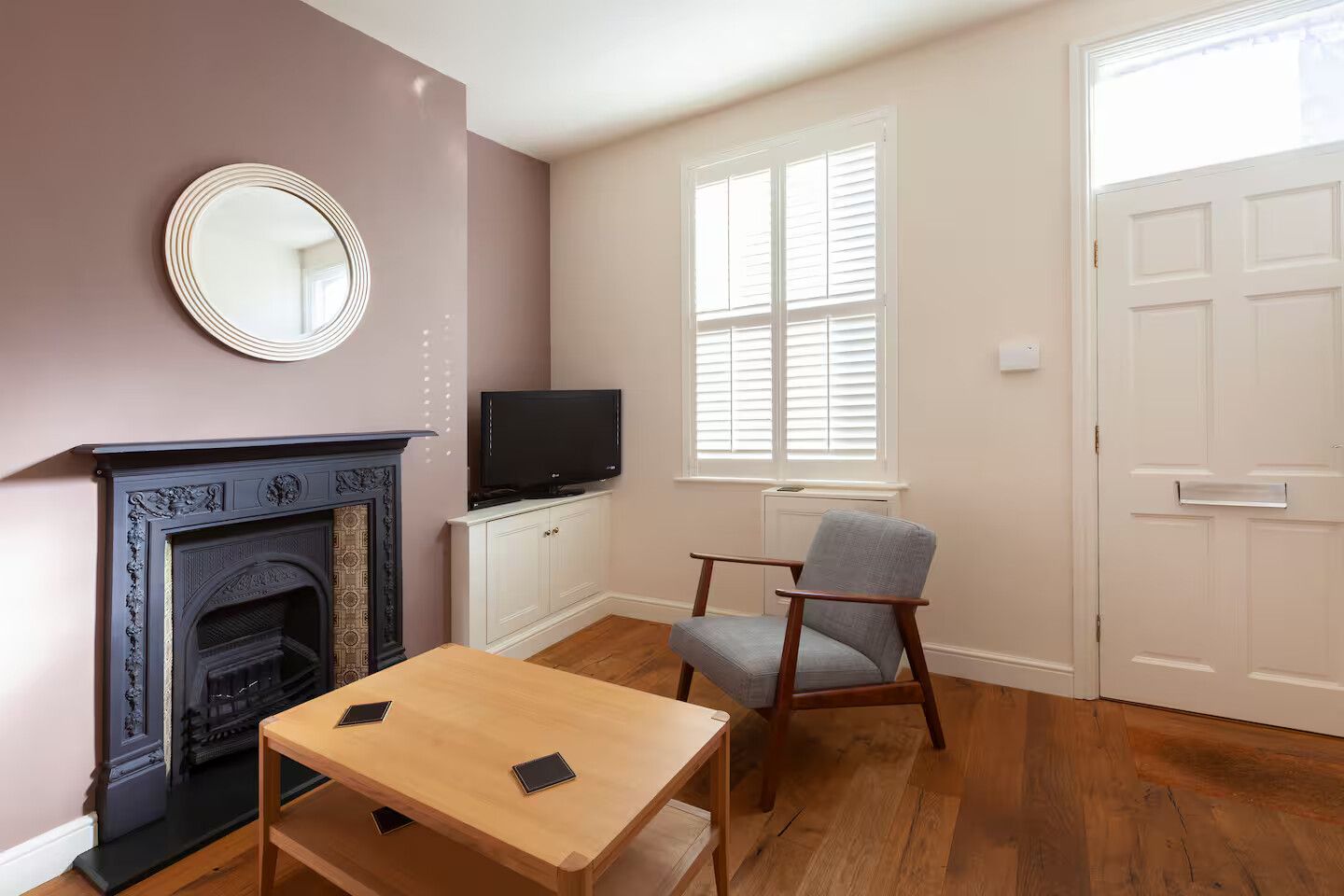 A room with a fireplace, a television on a cabinet, a table with two electronic devices, a chair, and a door.