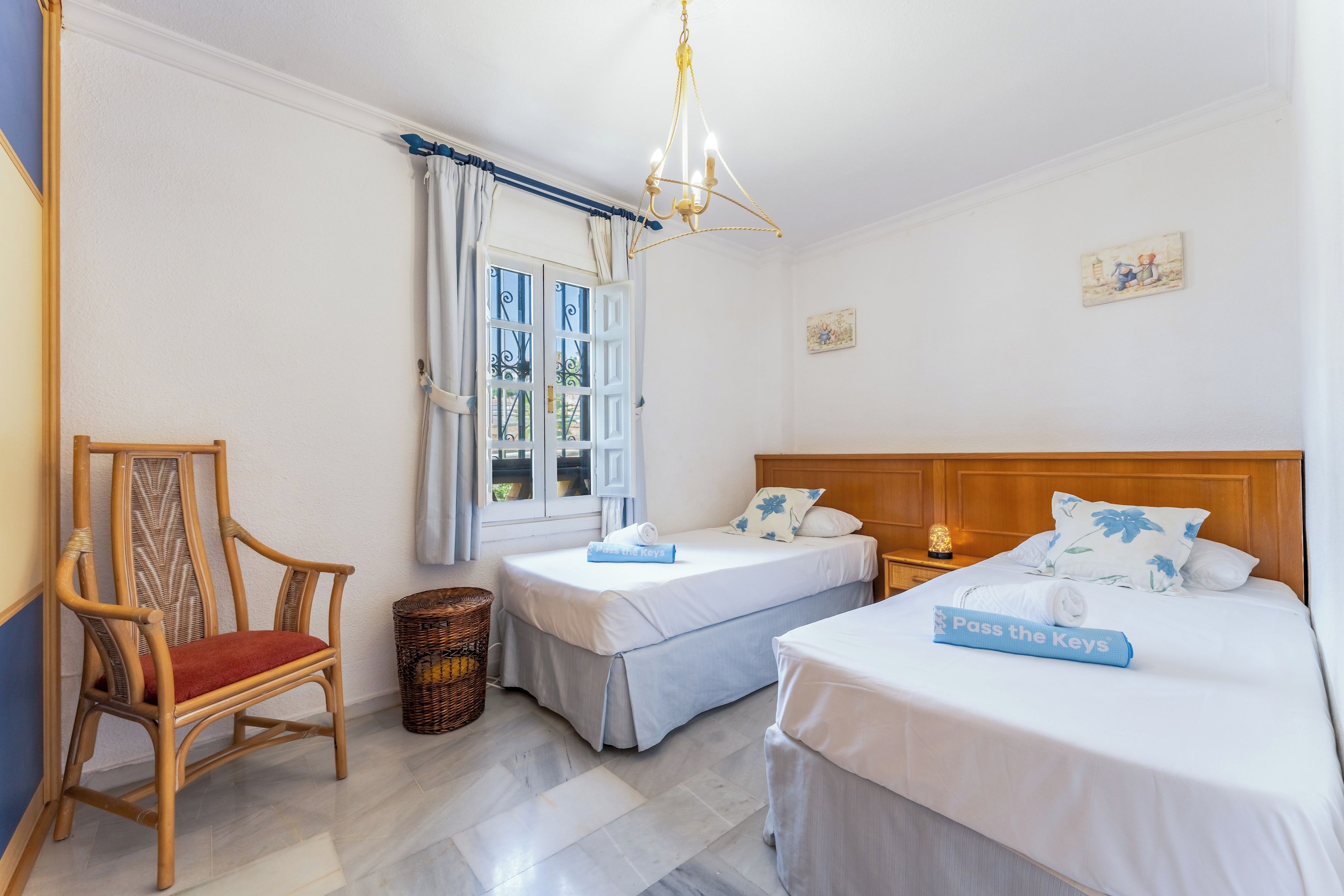 Quarto de hotel com duas camas de solteiro, uma cadeira de madeira, um cesto de vime e uma janela com cortinas.