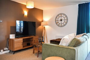 A modern living room with a flat-screen TV, wooden entertainment unit, floor lamp, and a green sofa. - Marina-View Flat near Cardiff & Penarth - Pass the Keys (Penarth)