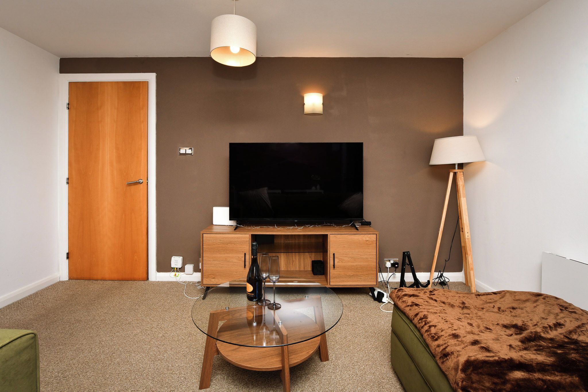 A modern hotel room with a flat-screen TV, a glass coffee table, a sofa, a wooden cabinet, and two floor lamps.