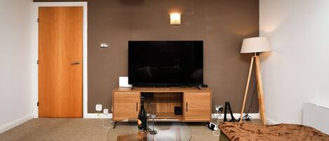 A modern hotel room with a flat-screen TV, a glass coffee table, a sofa, a wooden cabinet, and two floor lamps.