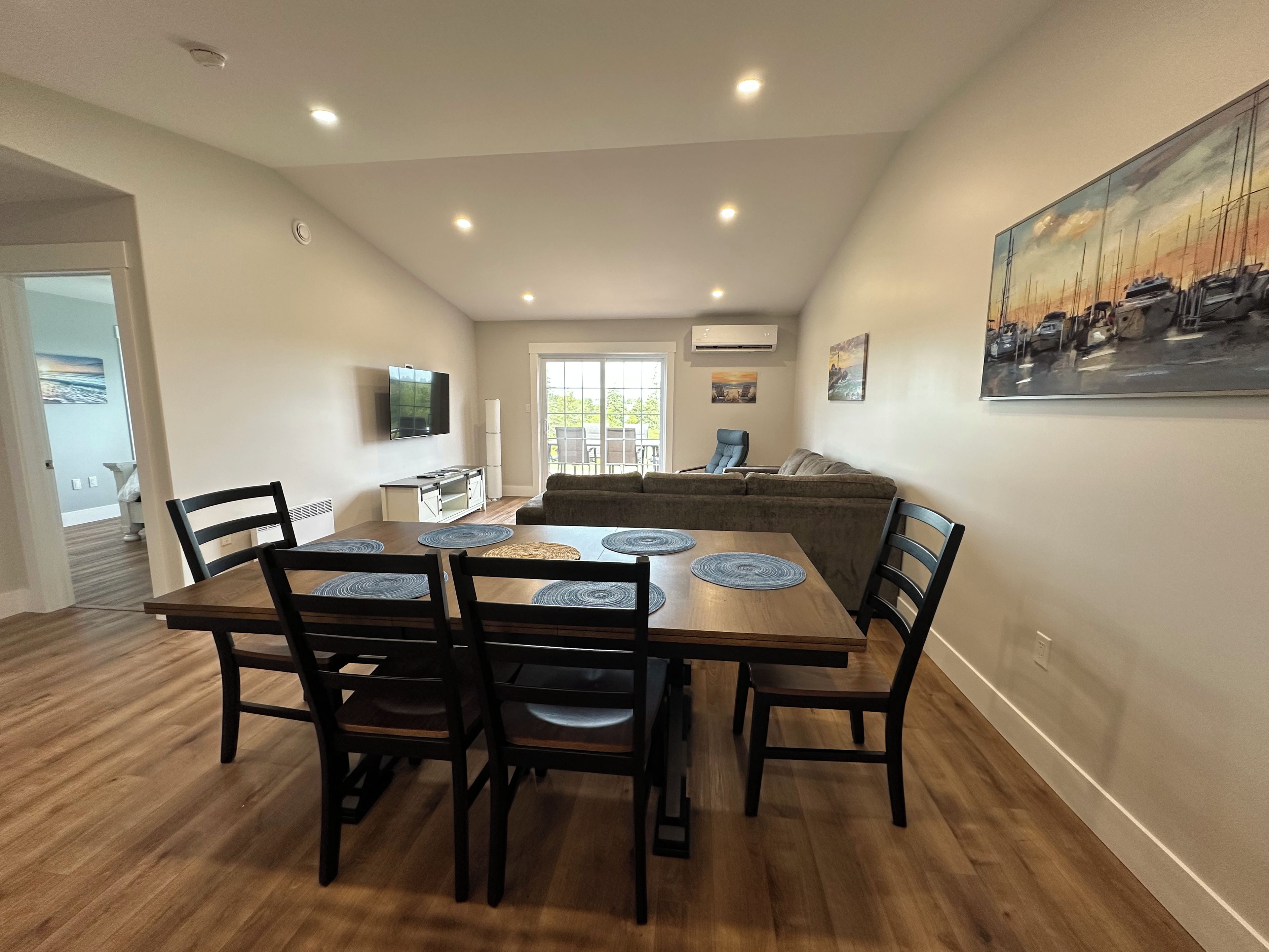 A dining area with a wooden table and chairs, a kitchen in the background, and a painting on the wall.