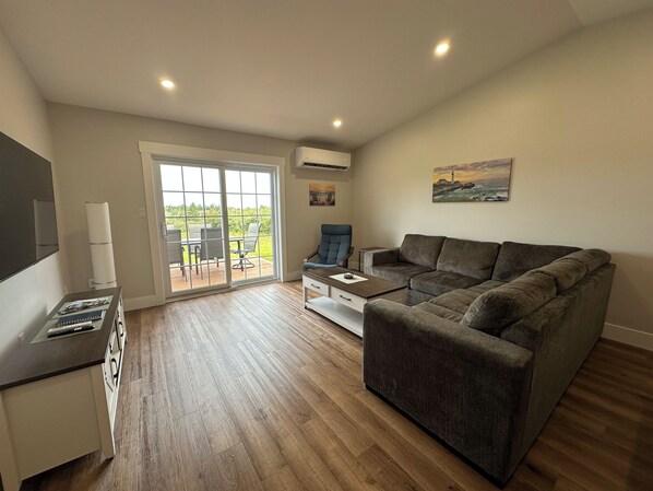 A spacious living room with a large grey sofa, a small table, and a TV on a stand. There is a sliding glass door leading to an outdoor area with a table and chairs.