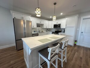 Una cocina moderna con armarios blancos, electrodomésticos de acero inoxidable, una isla central con taburetes de bar, iluminación colgante y suelo de madera.