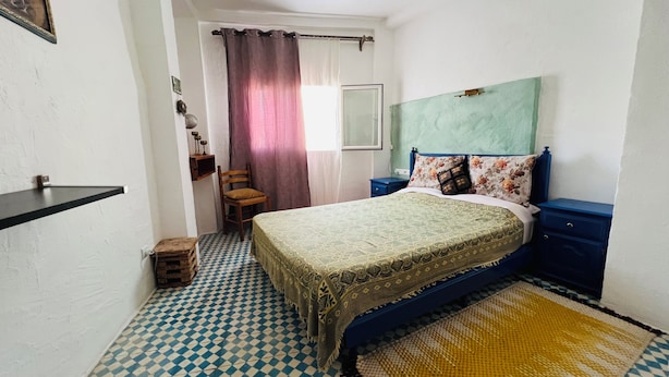 A bedroom with a checkered floor, a bed with a patterned bedspread, a chair, a nightstand, and a window with curtains. - Maisonette with Rooftop Terrace at Bab Souk – AC, WiFi & Medina Views (Chefchaouen)