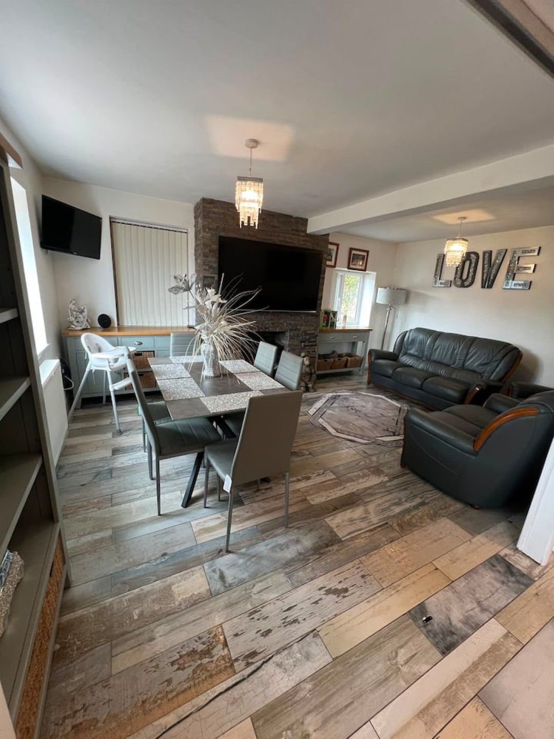 A modern living room with a fireplace, a dining area with a table and chairs, a black leather sofa, and a TV mounted on the wall.