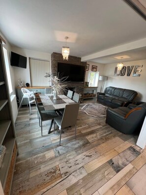 A modern living room with a fireplace, a dining area with a table and chairs, a black leather sofa, and a TV mounted on the wall. - Entire Executive home singles, couples, groups. (Bridgend)
