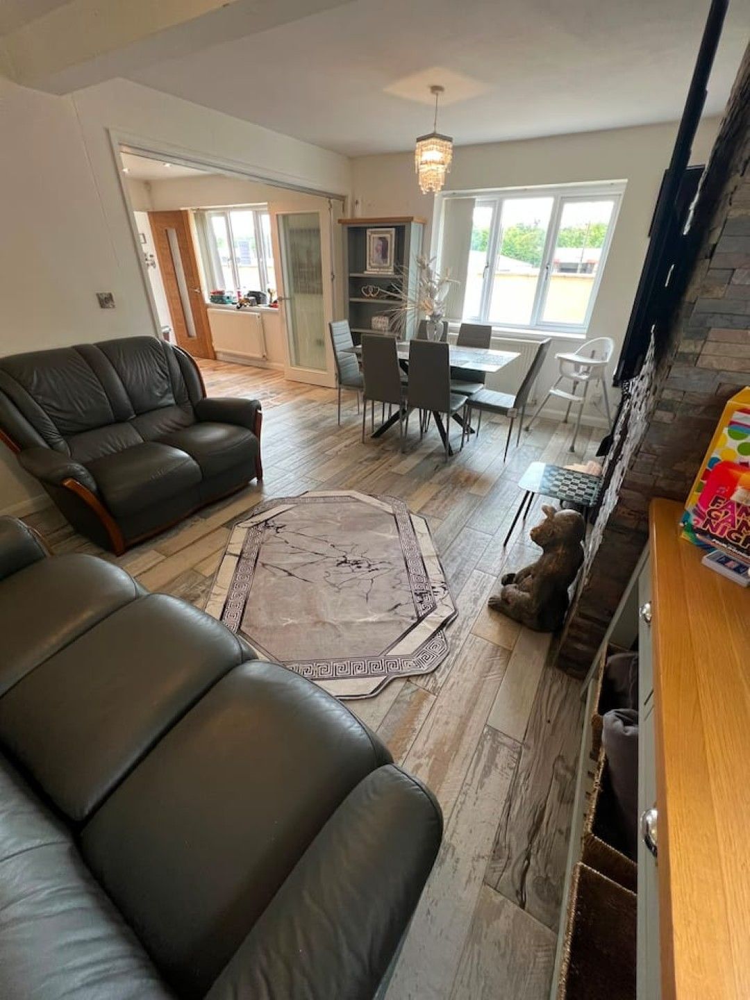 A living room with a black leather sofa, a dining table with chairs, a rug, and a teddy bear on the floor.
