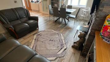A living room with a black leather sofa, a dining table with chairs, a rug, and a teddy bear on the floor.