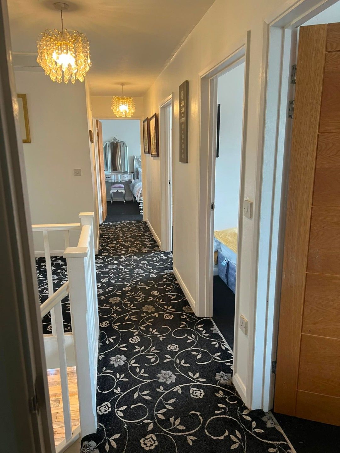 A hallway with a patterned carpet, a chandelier, and a bed with a white headboard in the background.