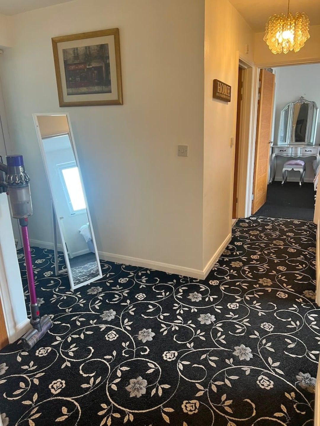 A hallway with a black floral carpet, a full-length mirror, a vacuum cleaner, and a vanity with a mirror.