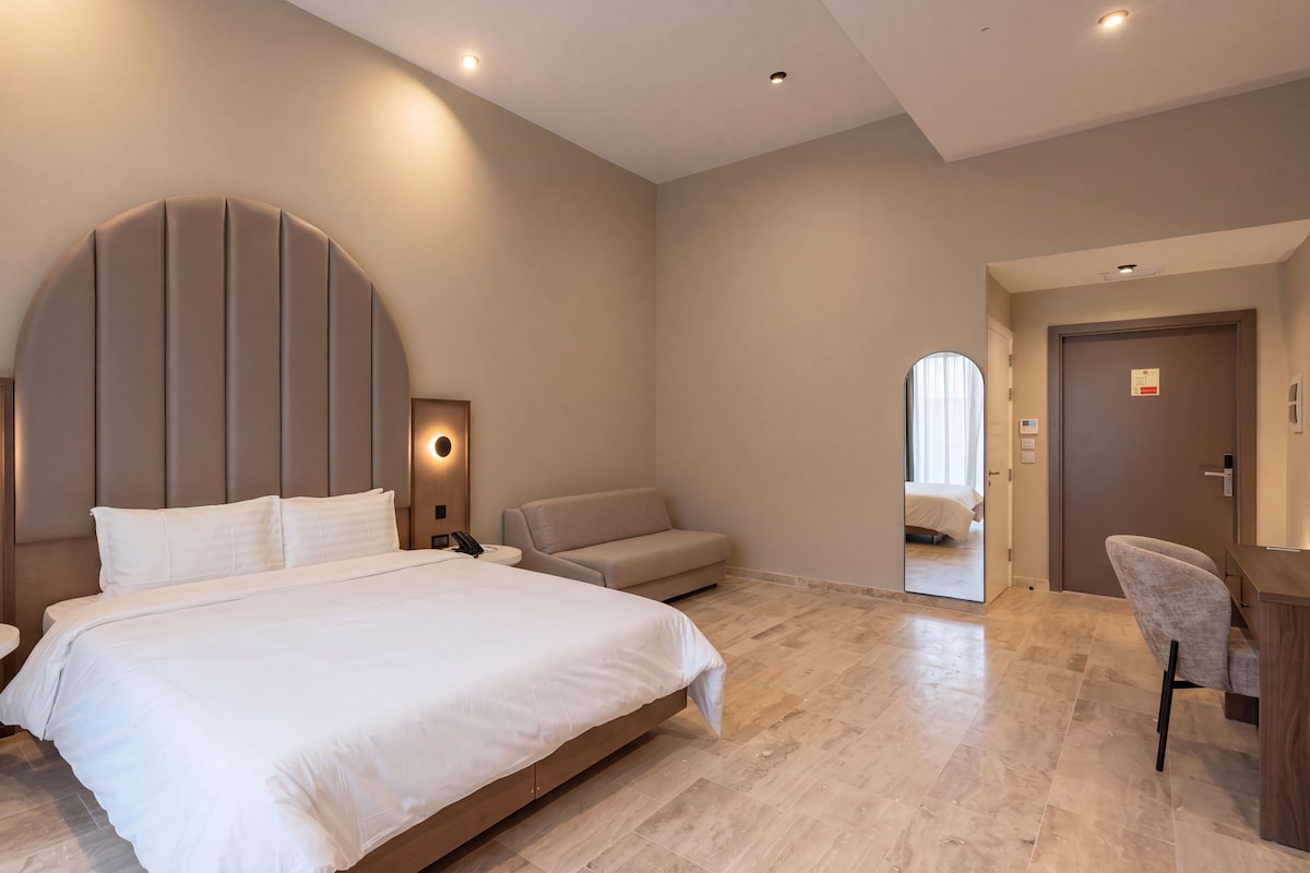 A modern hotel room with a large bed, bedside tables, a sofa, and a desk.