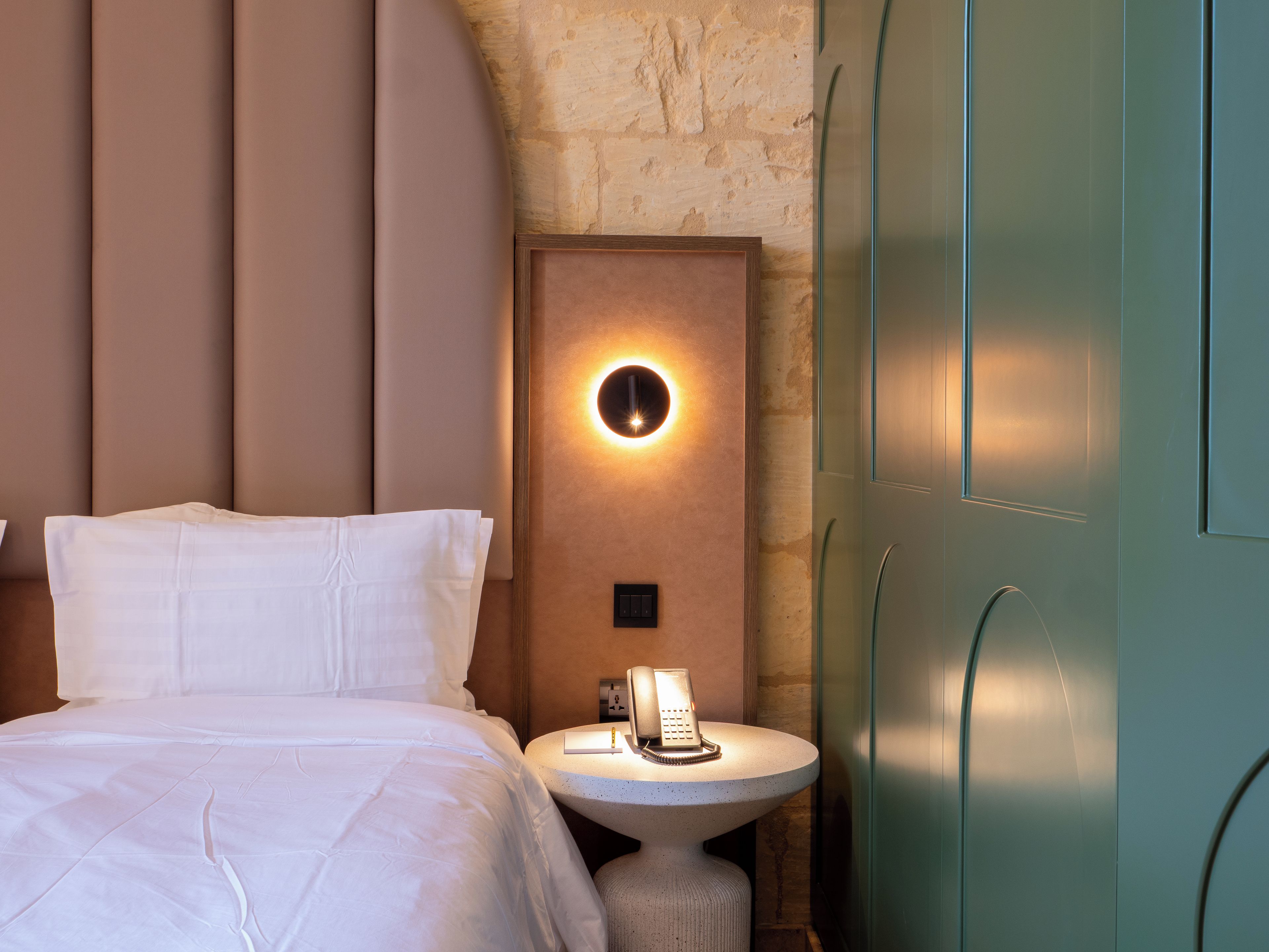 A hotel room with a bed, a bedside table with a lamp, a telephone, and a wall-mounted light fixture.