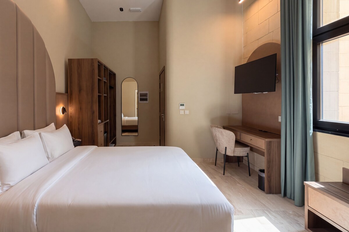 A hotel room with a large bed, a desk, a chair, a TV, and a wardrobe.