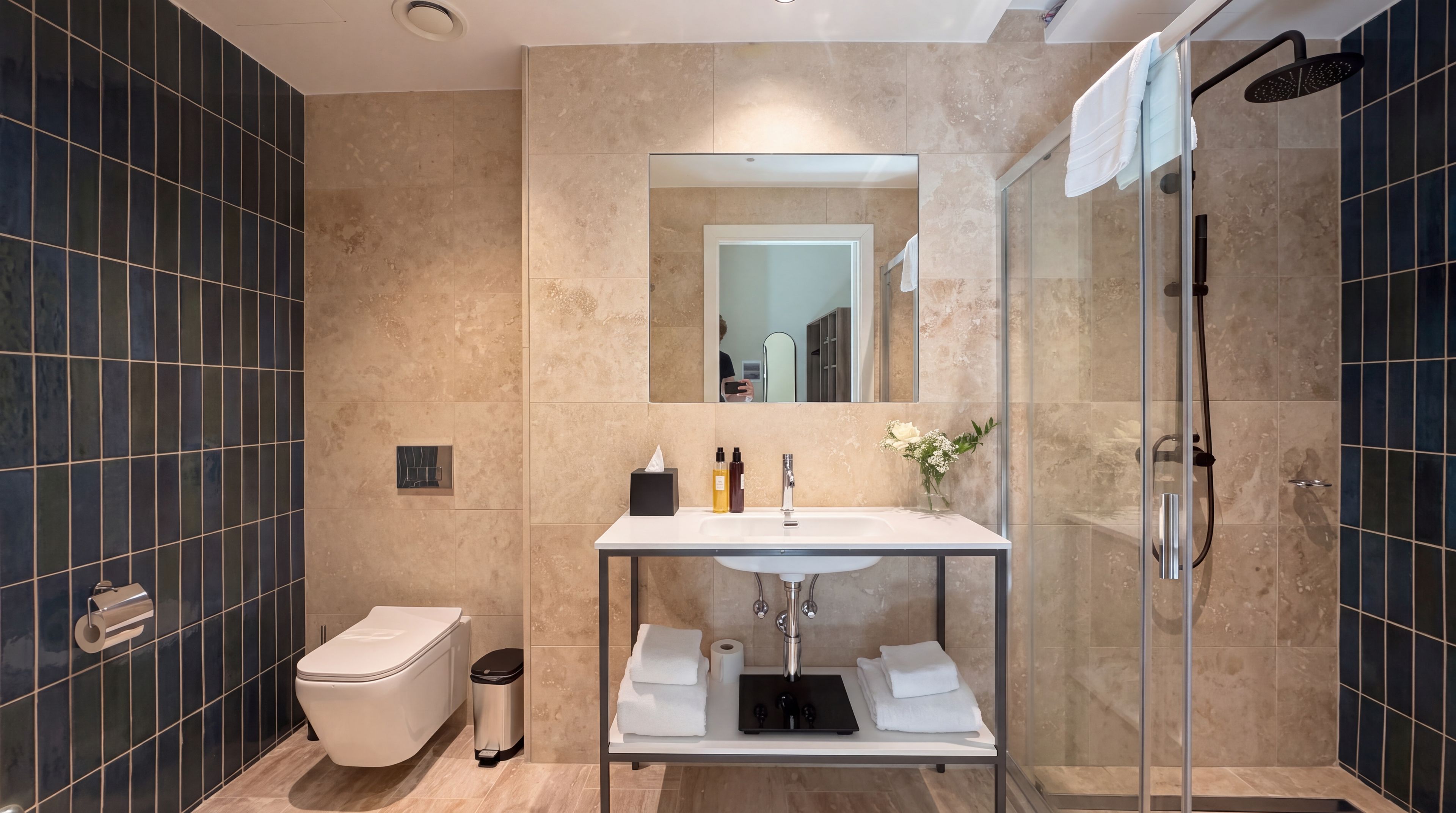 Une salle de bain moderne avec une douche  à parois de verre, un lavabo blanc avec un miroir au-dessus et des toilettes avec une poubelle.