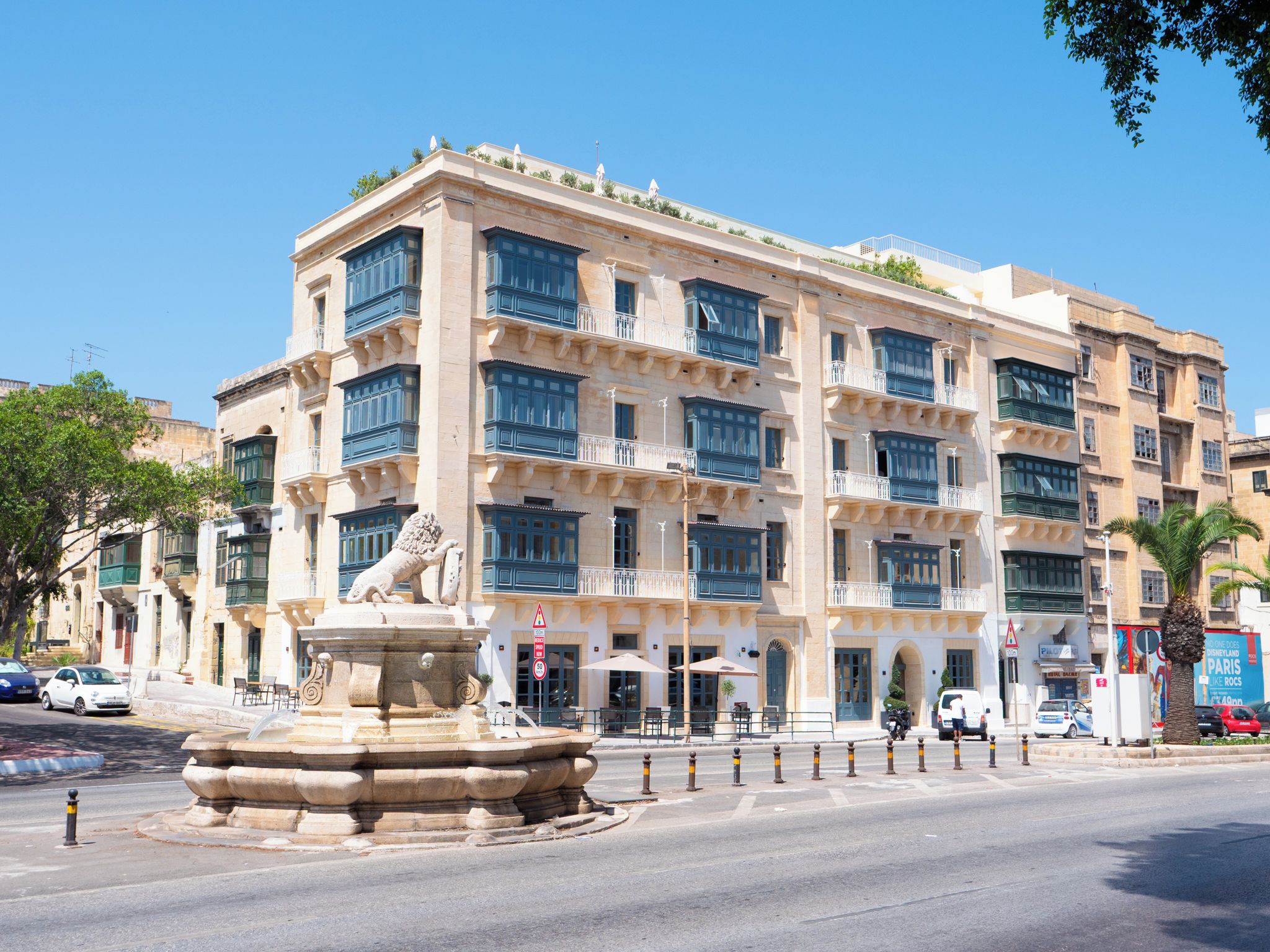 A multi-story building with a central courtyard, a fountain with a lion statue, and a street with parked cars.