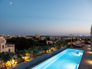 Una piscina en la azotea con vistas a la ciudad, sillas de descanso y una zona de jardín bien iluminada.