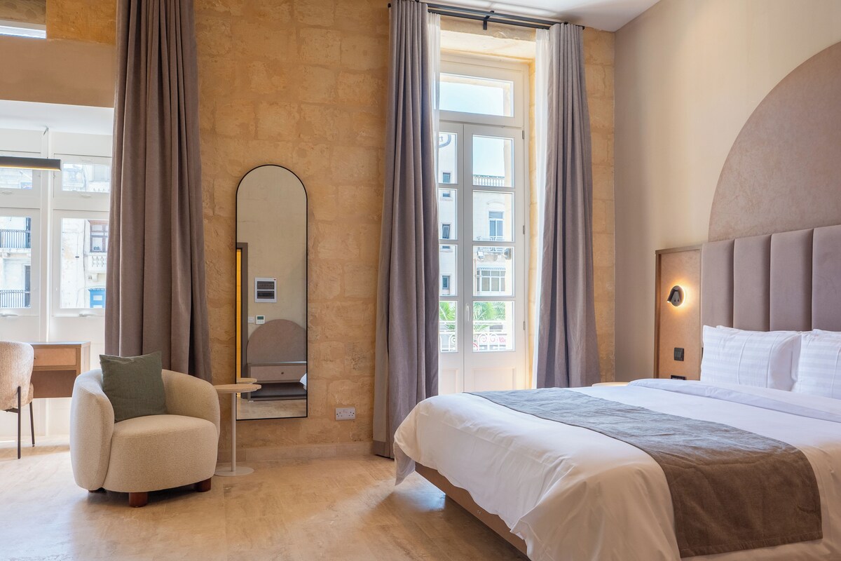 A hotel room with a bed, a chair, a round mirror, and a view of buildings through the window.