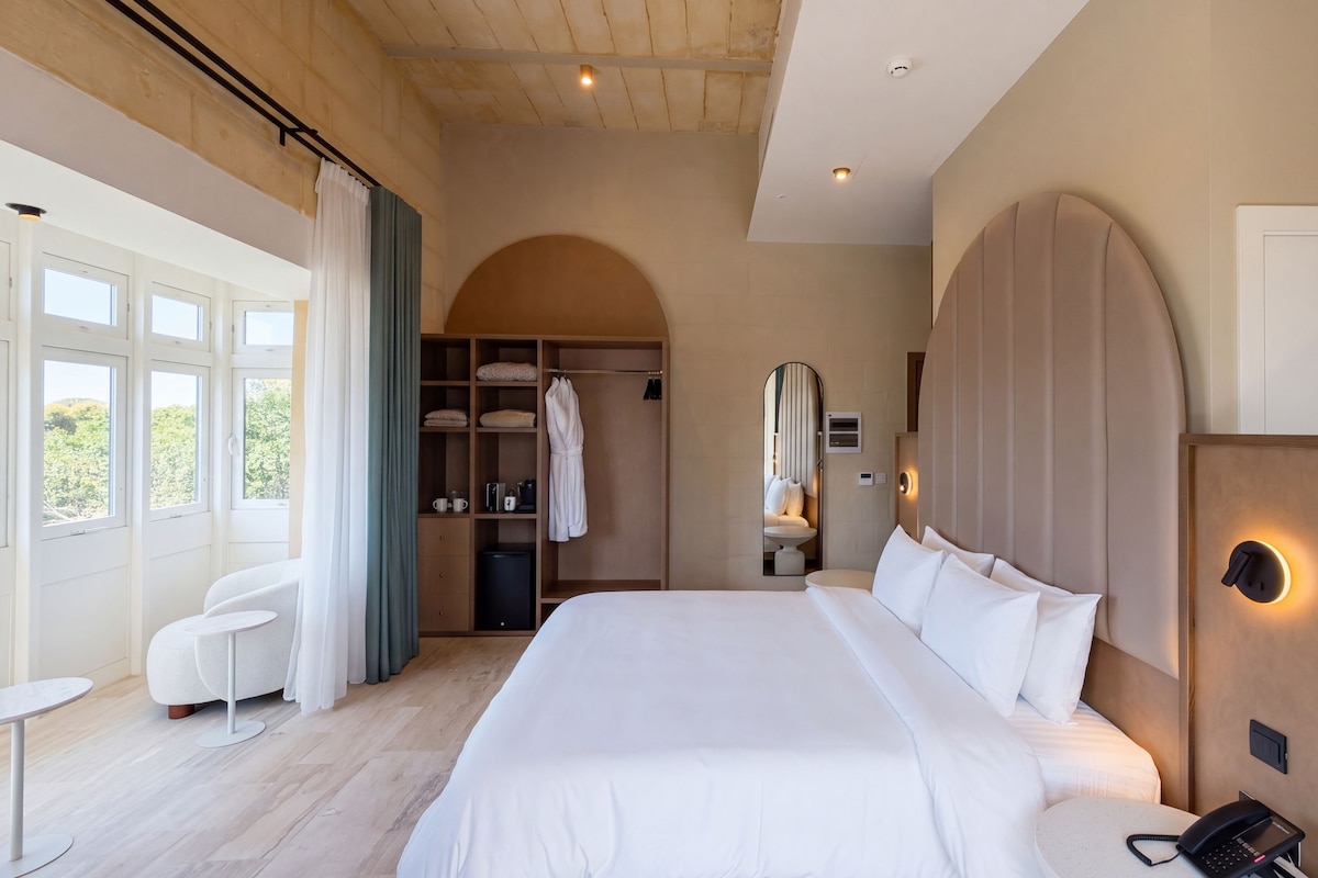 A hotel room with a large bed, a seating area with a table, a wardrobe, and a bathroom with a mirror.