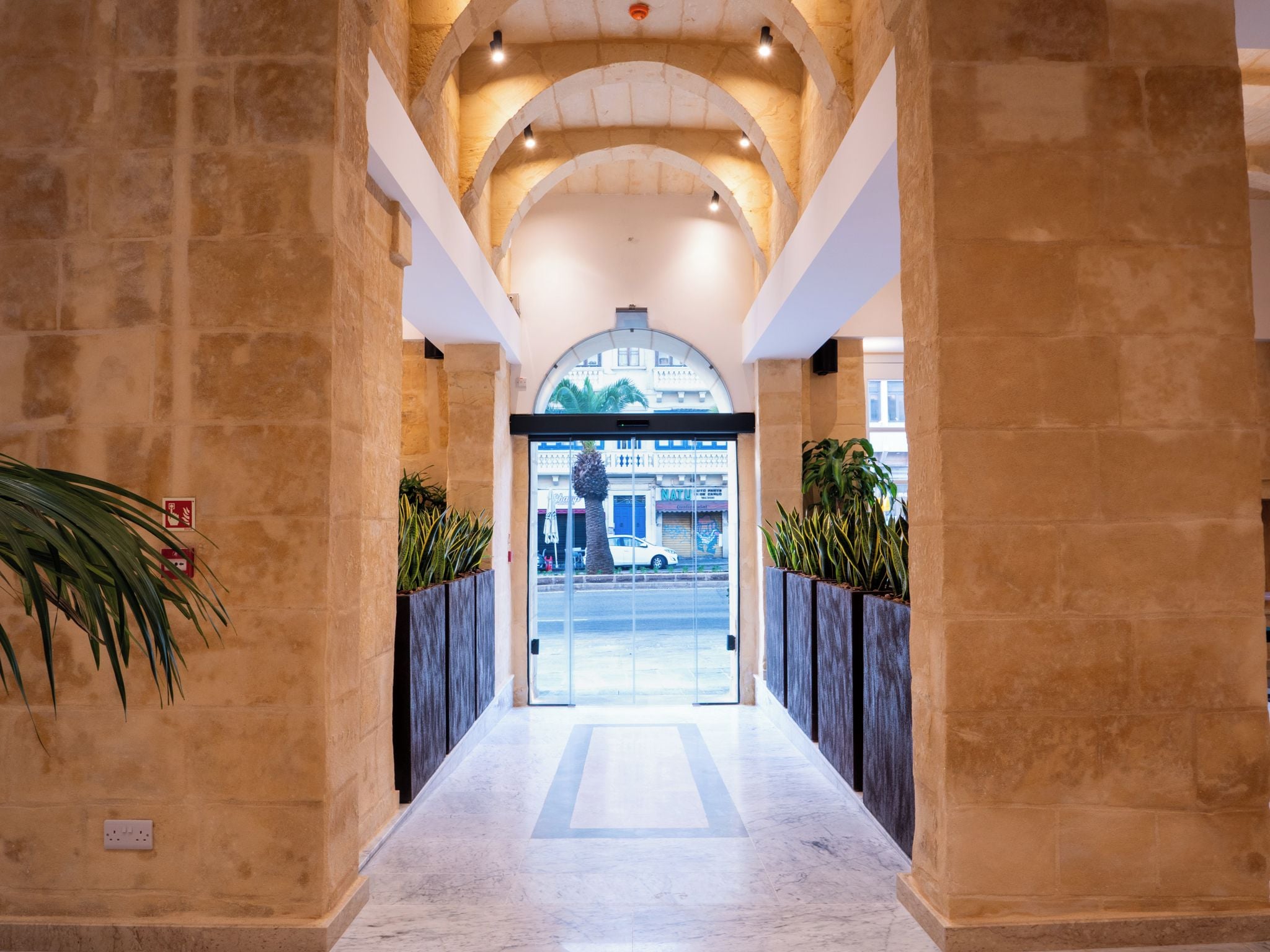Un pasillo con paredes de piedra, un acceso por puerta de cristal y plantas en macetas.