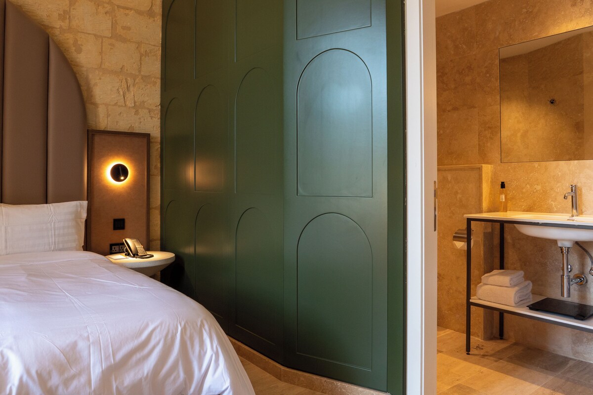 A hotel room with a bed, a bedside table with a lamp, a green accent wall, and a bathroom with a sink and towel rack.