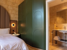 Quarto de hotel com cama, mesa de cabeceira com abajur, parede verde decorativa e banheiro com pia e porta-toalhas.
